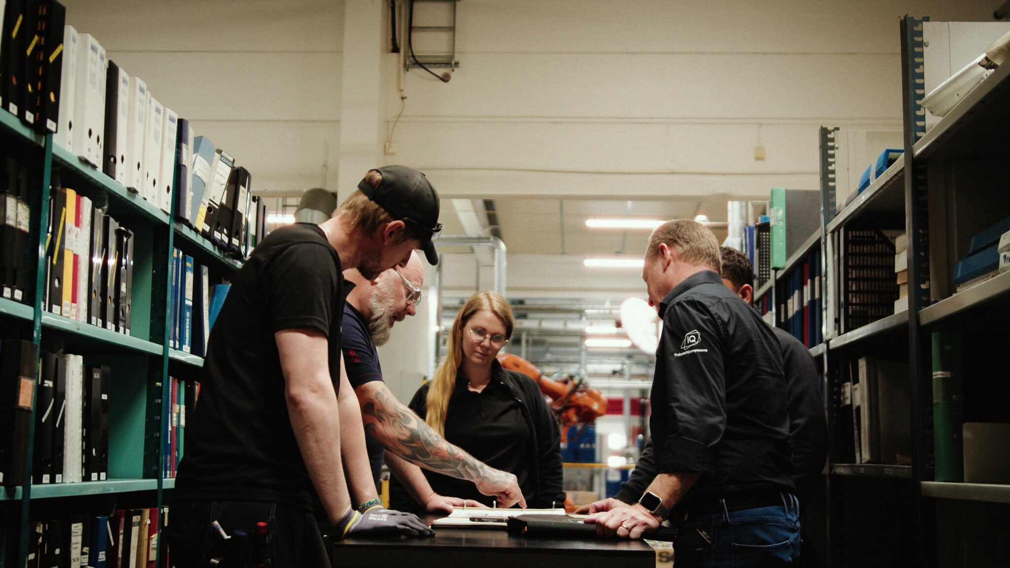 Medarbetare inom industrin står samlade vid ett arbetsbord i en fabriksmiljö och går igenom dokument mellan hyllor med pärmar och teknisk utrustning.